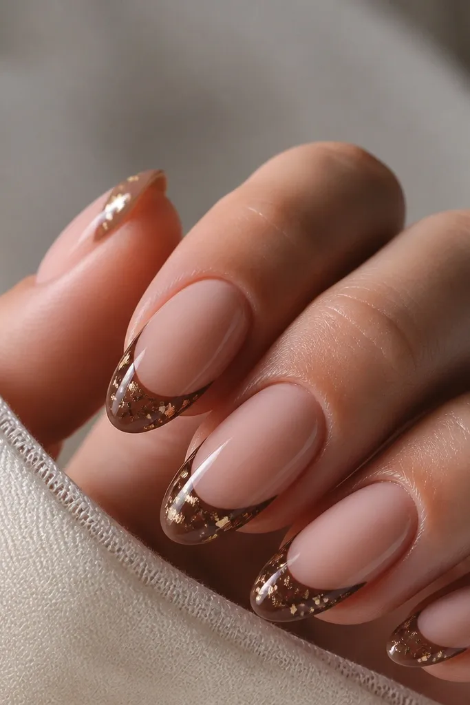 Nails with nude base and chocolate-brown French tips sprinkled with tiny gold foil flecks, glossy finish.
