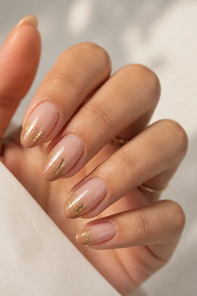 Oval nails with champagne nude ombre; a few irregular gold foil shards sit on the side of the mid-nail, and the tip fades into a slightly deeper nude.