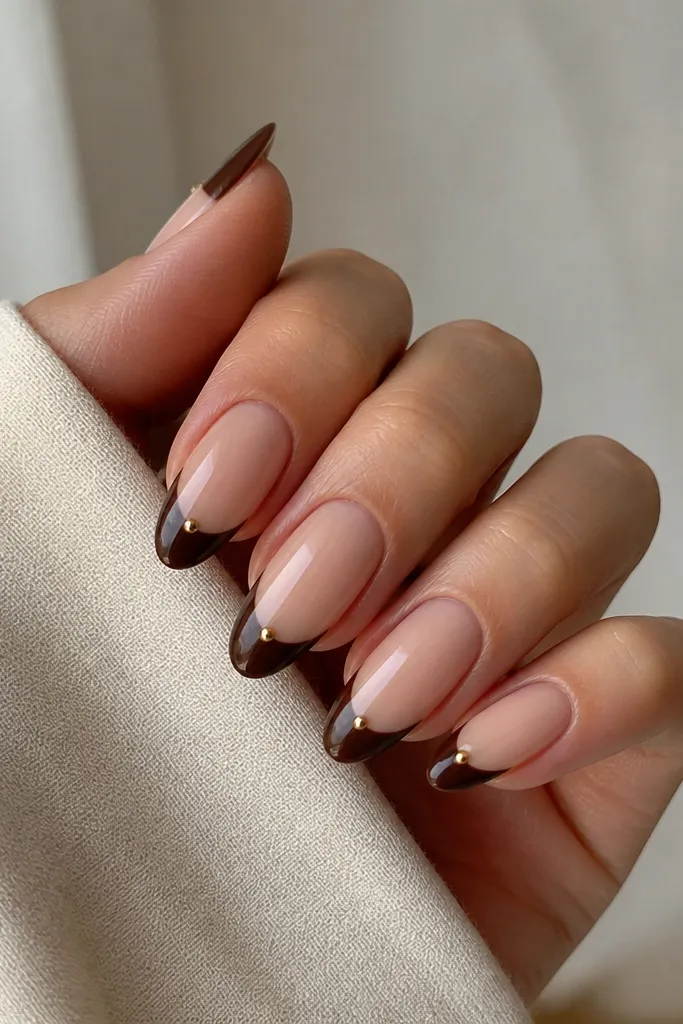 Almond nails with a nude base and glossy brown French tips. Each tip has a tiny metallic gold dot near one corner of the smile line.