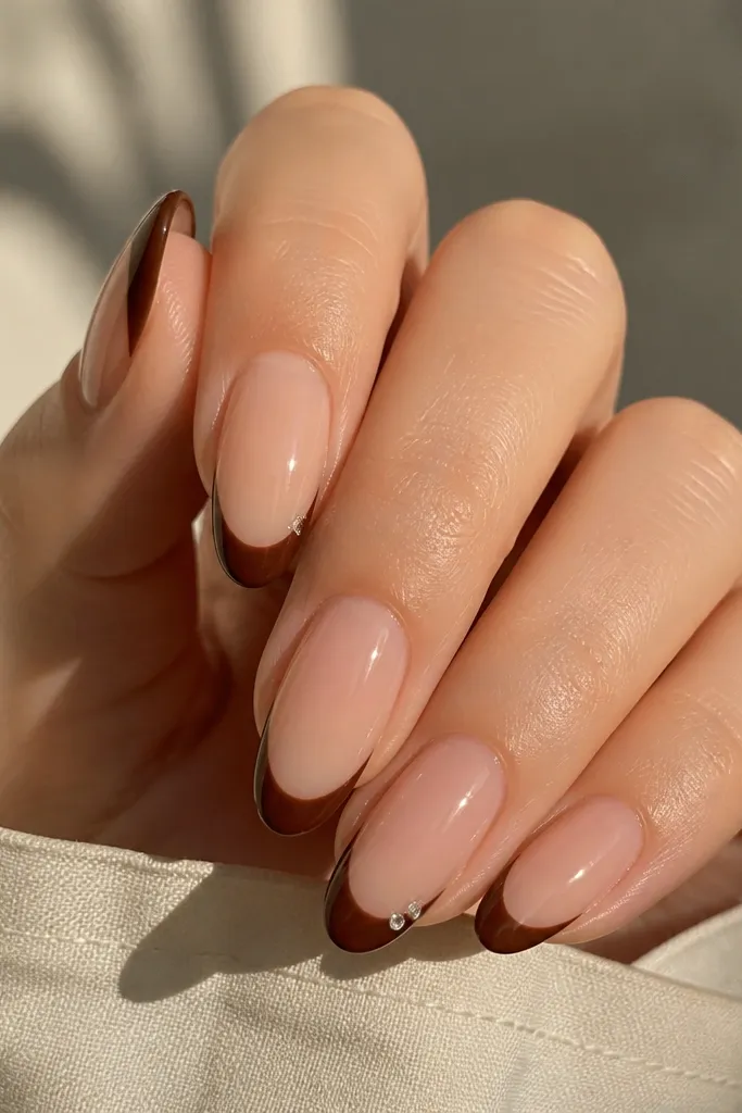 Five almond nails. Sheer nude base. Chocolate brown french tips with a glossy finish. On two nails, a single tiny clear rhinestone sits near the outer corner of the tip, set into the brown.