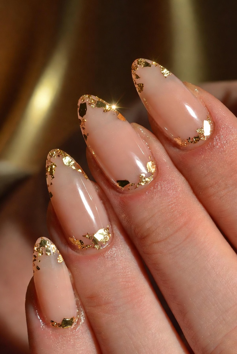 Long coffin nails in sheer nude. At the cuticle there are irregular gold foil flecks, like tiny broken leaf pieces, with a glossy finish.