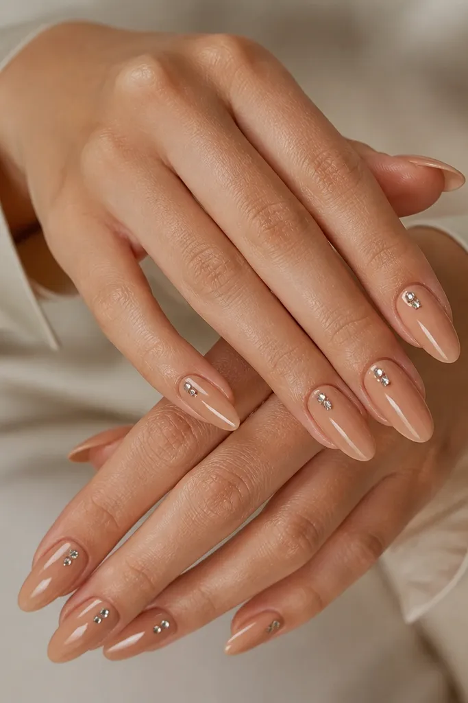 Long almond nails in caramel nude. On each nail there is a cluster of three tiny rhinestone studs near the cuticle, glossy top coat.