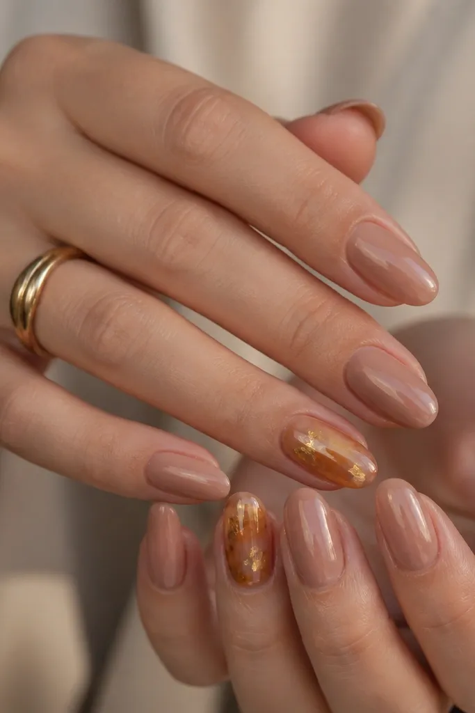 Warm nude nails with two accent nails coated in a translucent amber glaze. Around the center of those accent nails are small gold leaf pieces scattered like warm light. Other nails stay nude and glossy.