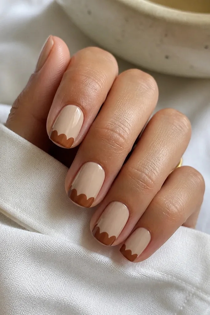 Short squoval nails with a warm beige base. The tips have scalloped arcs in cinnamon brown, creating a repeating half-circle pattern across the edge. The scallops are evenly sized and glossy.