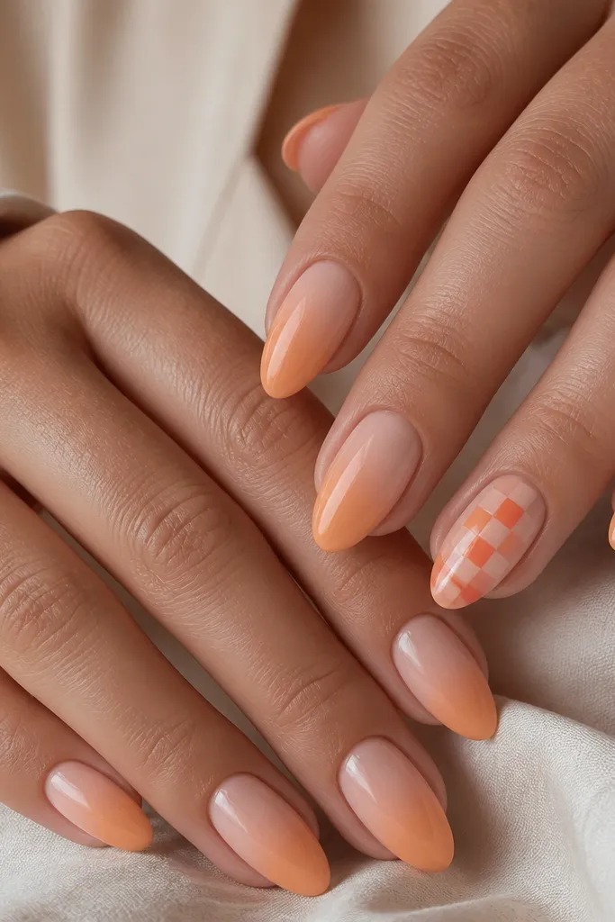 Nude-peach nails with a subtle orange gradient on most nails; one accent nail per hand has a tiny 2x3 checker pattern in coral and peach along the center; glossy topcoat.