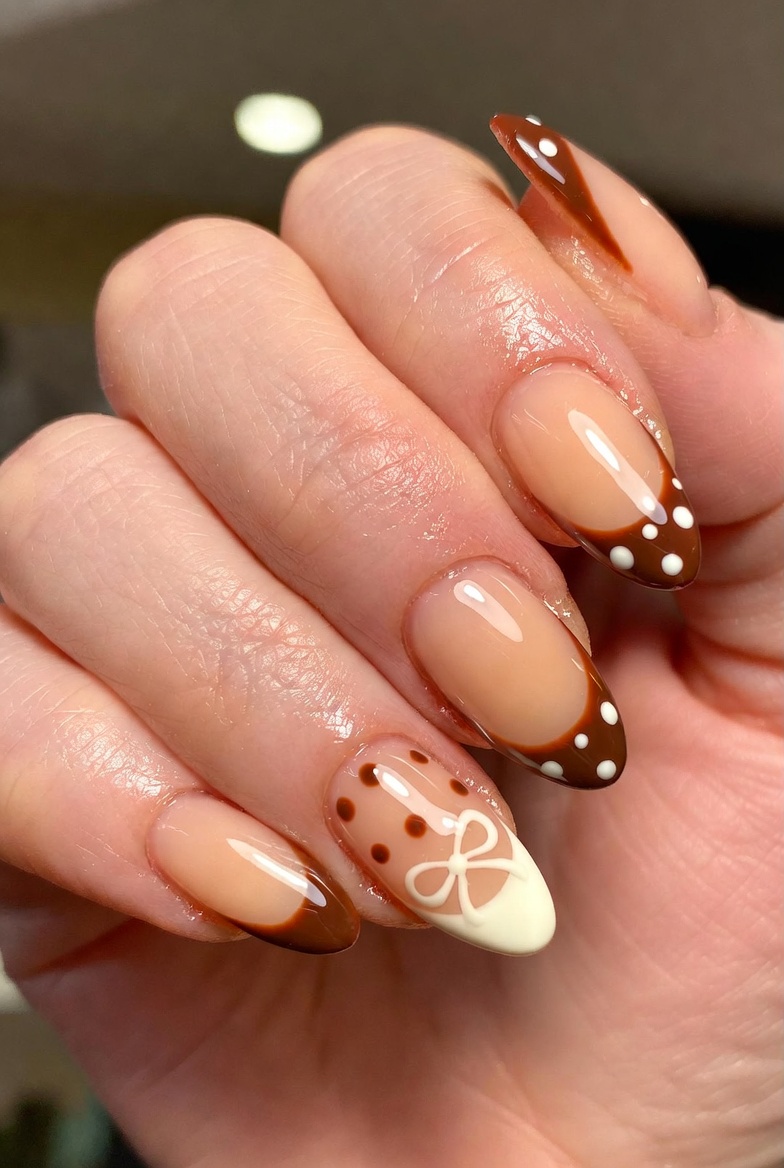 Glossy brown French tips on a sheer nude base. Instead of single dots, there are clusters: one larger dot, two smaller dots, and a micro dot, repeated near the center of each tip.
