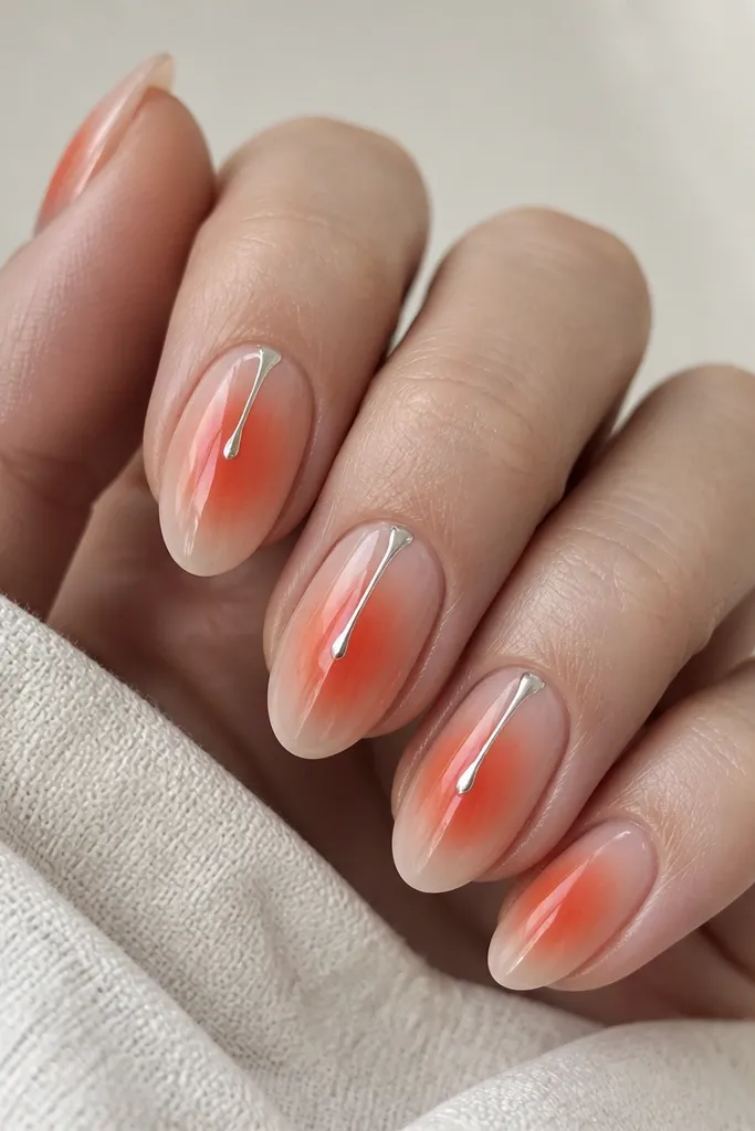 Nails with a warm orange-coral aura centered and fading outward. On two nails, a thin reflective silver drip hangs from the top third down toward the aura center.