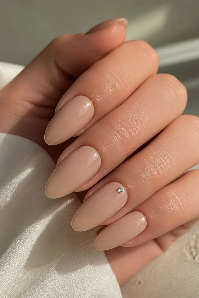 Almond nails in neutral beige nude; one nail has a single tiny rhinestone placed near the cuticle center; glossy top coat.