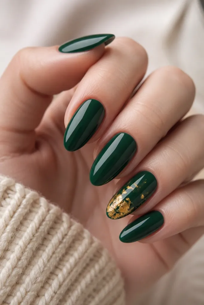 Most nails are emerald glossy; one accent nail has irregular gold foil leaf fragments clustered near the tip, with some foil edges feathering outward.