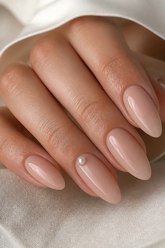 Almond nude nails in a sheer pink-beige tone. One nail has a single tiny 3D pearl dot right near the cuticle area; the rest are plain glossy nude. The pearl looks like a small teardrop pearl bead.