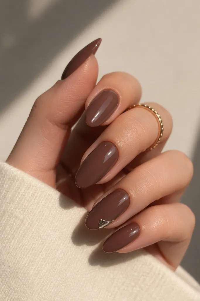 Almond nails in a deep cocoa nude. On one nail, a small silver foil corner sits near the free edge on one side, like a metallic crescent. Everything is glossy and smooth.