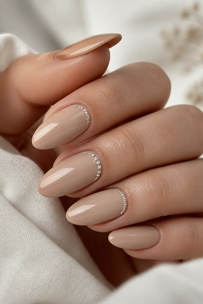 Almond nude nails in a neutral beige. A thin line of micro rhinestones runs along the cuticle curve on one or two nails. The rhinestones are tiny, evenly spaced, and the rest is plain glossy nude.