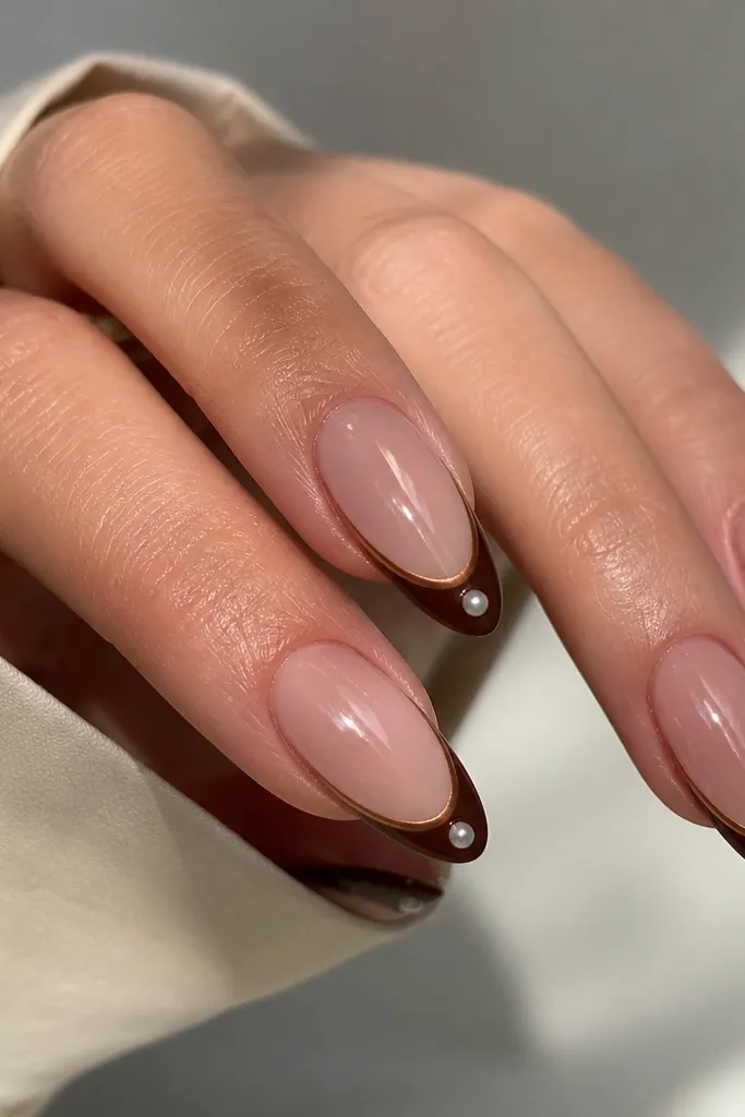 Almond nails with a deep cocoa French tip. There's a thin line of brown chrome along the outer edge, plus a single tiny pearl on the center of each tip. Glossy, with pearl highlights catching light.