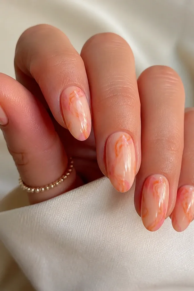 Medium oval nails. Pale peach base with swirling burnt orange marble veins and tiny touches of pink near the edges. Finish is glossy and glassy.