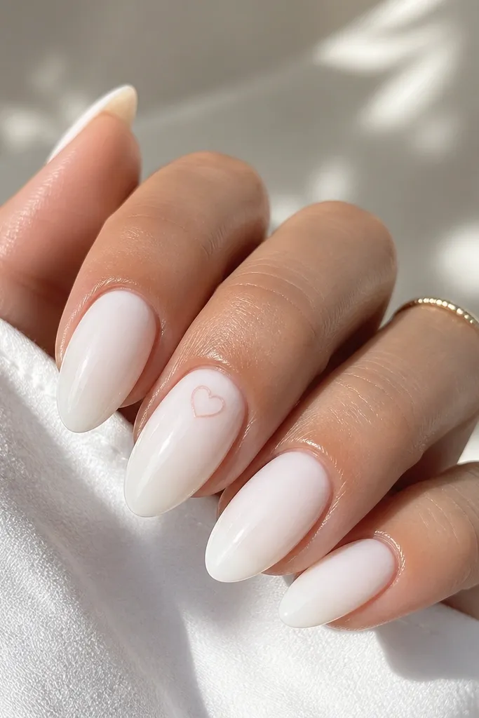 Milky white almond nails with one accent nail featuring a small thin-outline heart near the cuticle area. The heart is outlined in soft pink or blush gel. Glossy top coat.