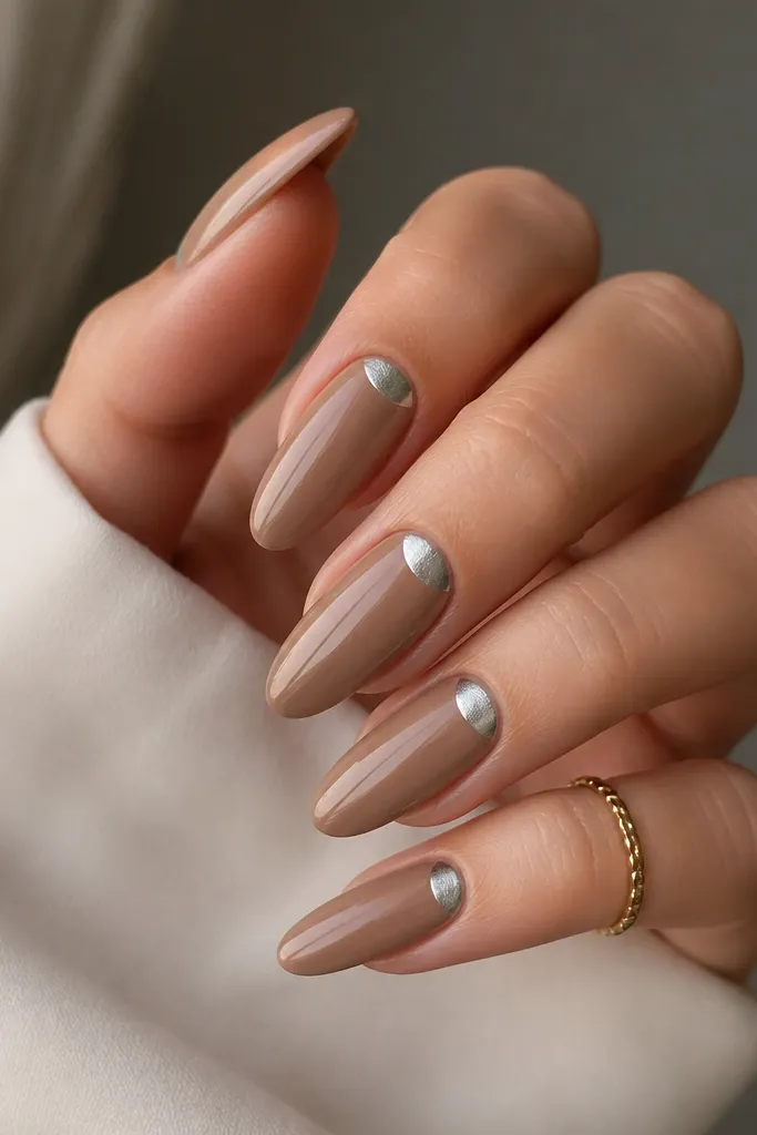 Long almond nails in cappuccino nude (light brown-beige). A frosted silver half-moon sits at the cuticle on each nail like a moon phase sticker, with glossy top on the rest.