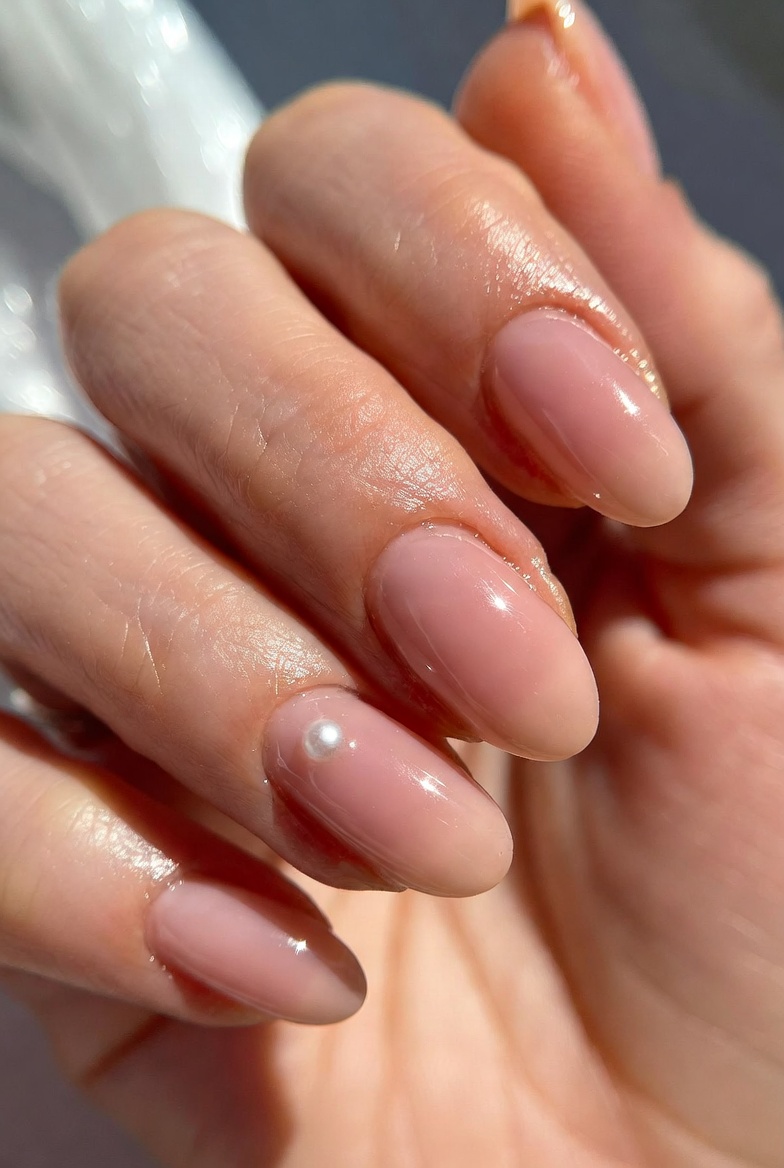 Short squoval nails in sheer ballet nude. The ring finger has one small round pearl at the center near the cuticle, with a glossy topcoat around it. Other nails are plain nude.
