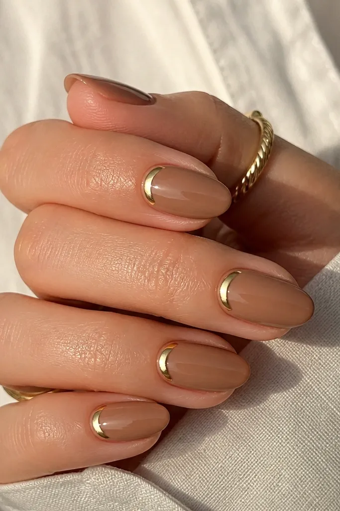 Short almond nails in caramel nude (warm beige with a hint of brown). On each nail, a small gold half-moon sits right at the cuticle. Glossy finish.