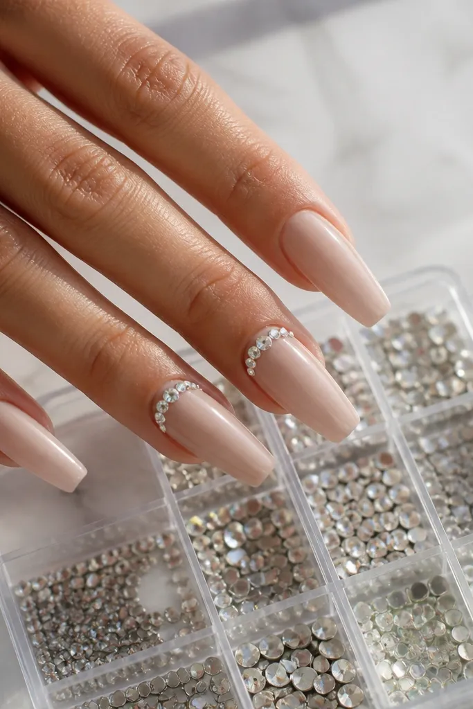 Nude coffin nails with a thin rhinestone arc near the free edge. The rest of the nails are glossy nude. A rhinestone storage box with grid compartments is shown.