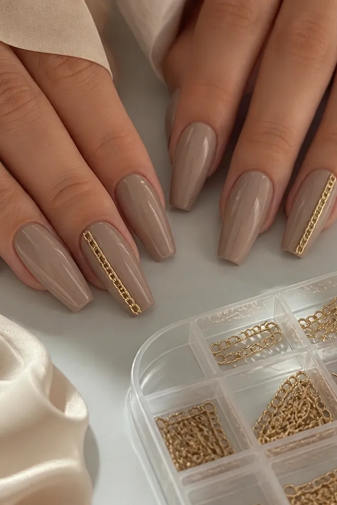 Taupe nude coffin nails with a single gold chain charm running along one side of the nail on two accent nails. A small charm chain storage compartment is visible.