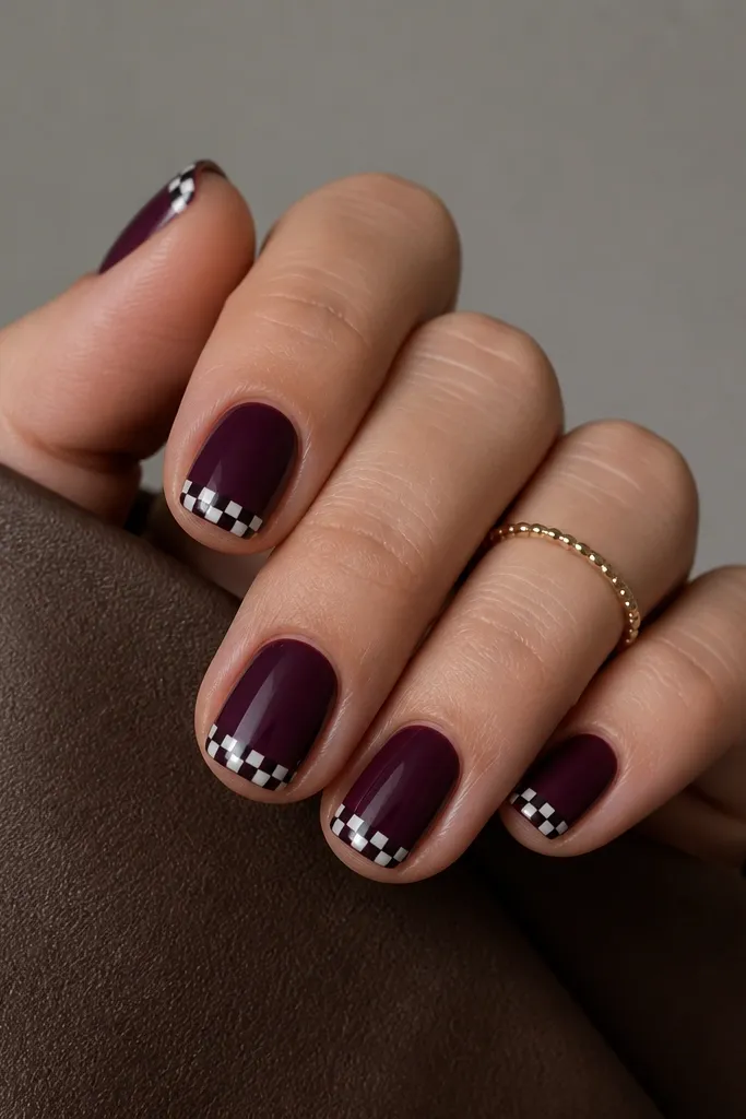 Short nails with a deep plum base. The tips have a small black-and-white checkered pattern made from tiny squares, about 3x3 squares per nail. The checkers sit only on the very tip area, leaving the rest solid plum.