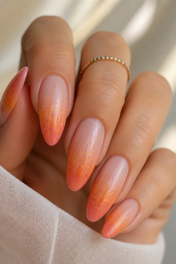 Almond nails with a sheer nude base and a soft sunset fade in the free edge. Tiny micro-glitter flecks in orange, pink, and gold are scattered lightly, mostly around the blend area. Glossy finish.
