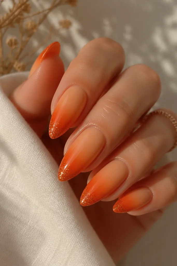 Almond nails with a gradient from peach nude to deep orange. The free edge has a sharper, darker orange fade and a light dusting of glitter only at the tips. Glossy finish.