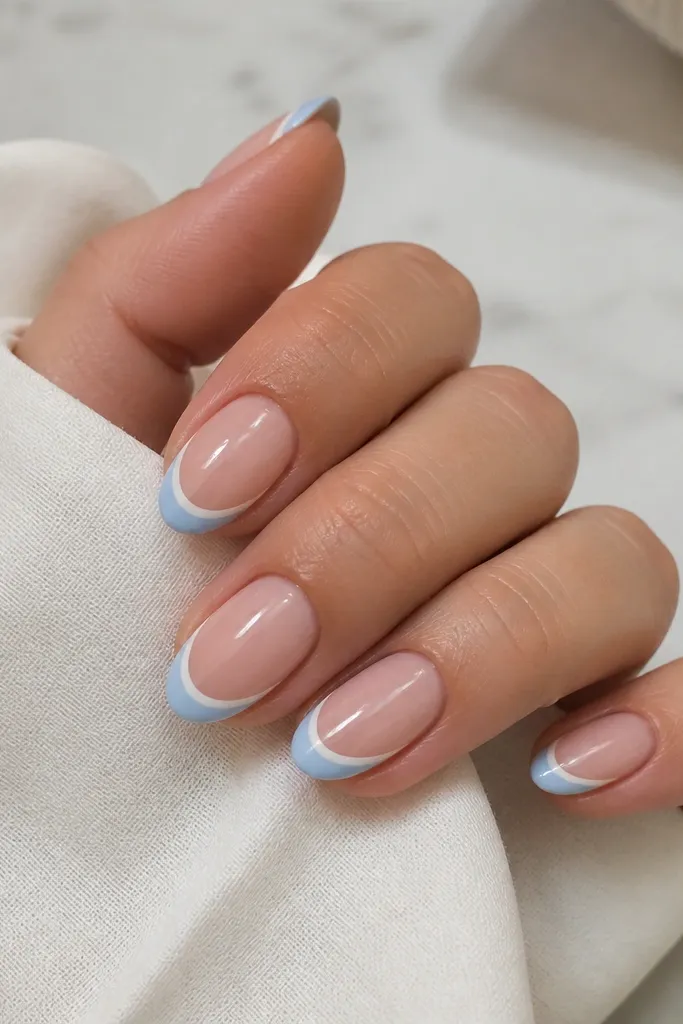 Short almond nails with nude-pink base. French tips are sky blue, and each tip has a hairline white line between the blue and the nude.