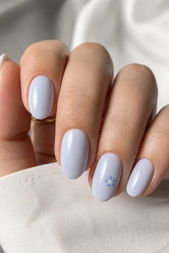 Short almond nails in a milky pale blue. On the ring finger only, a cluster of tiny sky blue dots forms a loose flower near the center of the nail, with one small white dot highlight.