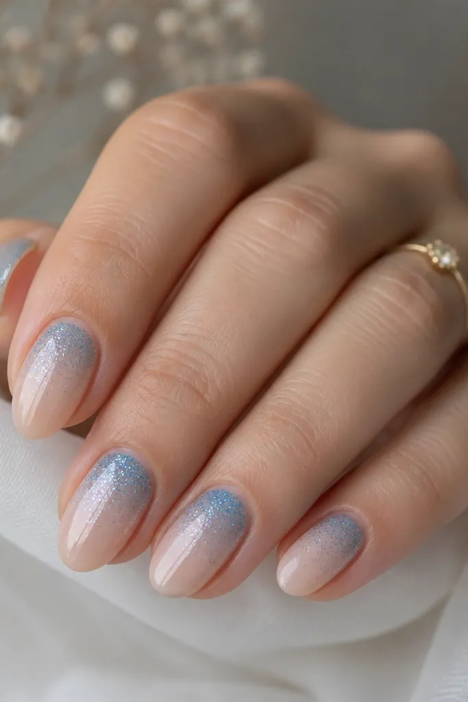 Short almond nails with a sheer nude base. The top third is a sky blue glitter fade, with the densest glitter at the very tip and fading downward.