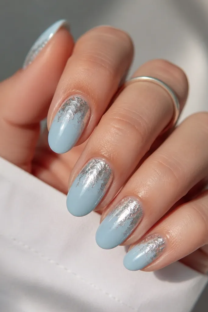 Short almond nails with a milky sky blue base. Half the nail near the cuticle is covered in irregular silver foil, with the foil edge blending into the blue.