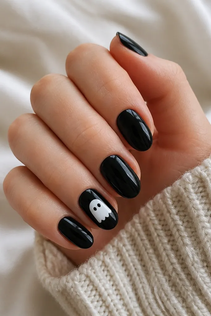 All nails are glossy black. The ring finger has a small white ghost face with two dot eyes and a simple rounded bottom. The ghost sits centered and does not touch the side edges.