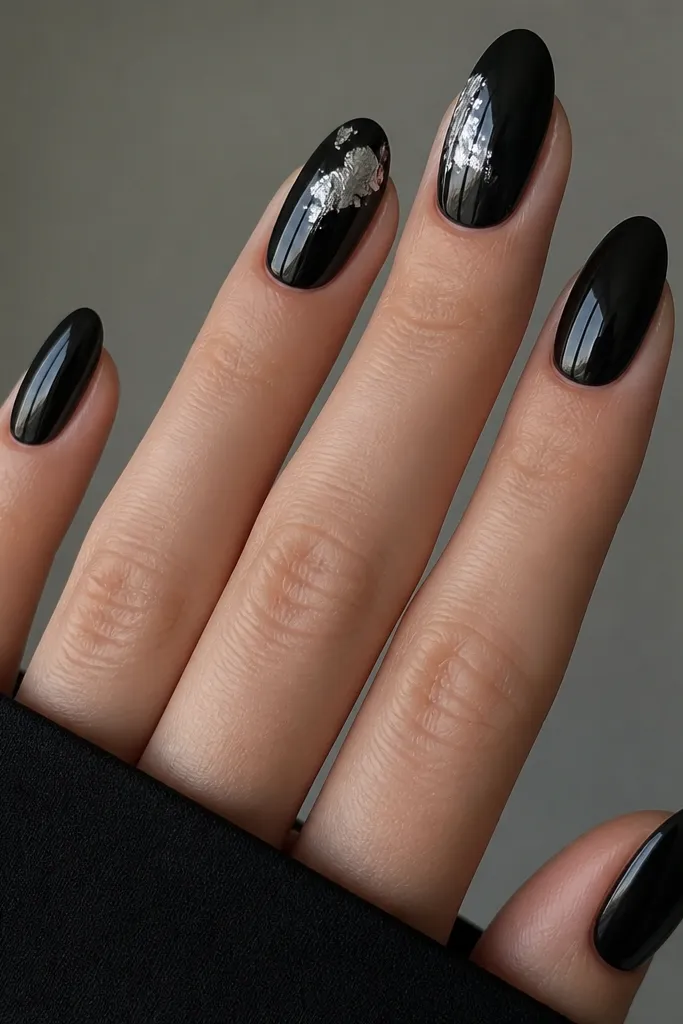 All nails are glossy black. On the index and middle fingers, small pieces of silver foil are placed like broken moon shapes near the upper half, with irregular edges and a few lines of foil reflecting light.