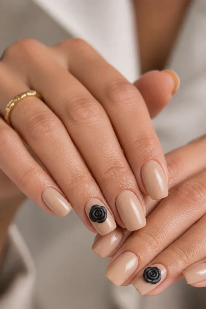 Opaque nude nails with glossy top coat. Two accent nails per hand have a black rosebud in the center: a small spiral bloom with thin petals. The petals have slight texture from layered black polish.