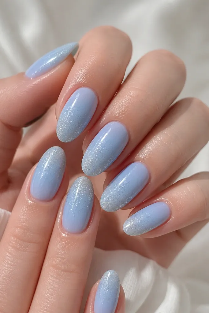 A full set of short almond acrylic nails in powdery sky blue. The tips fade into a fine, icy micro-glitter that looks like it's suspended in the clear coat. Cuticles are clean and glossy.