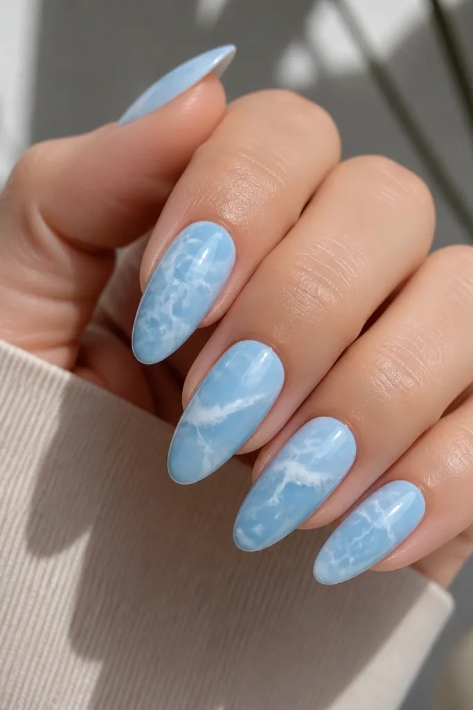 Almond acrylic nails in sky blue with swirling, milky-white marble veins. The veins look like thin cracks filled with opaque white, with a glossy finish.