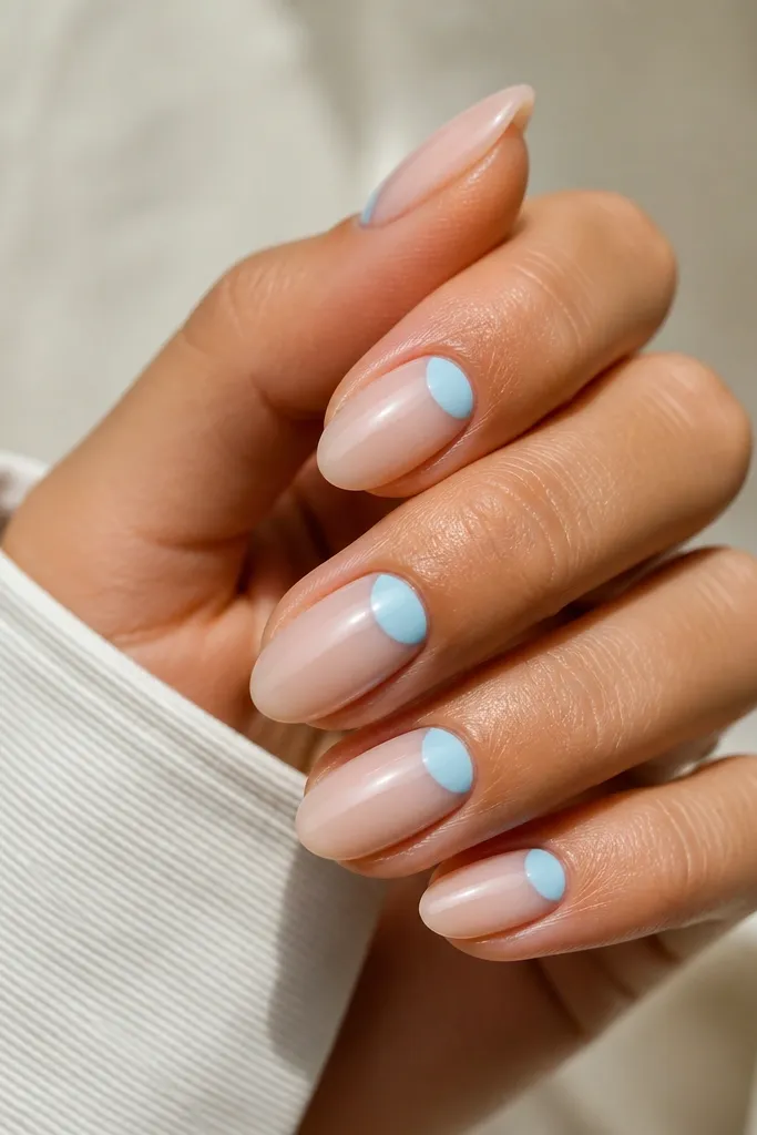 Medium almond nails with sheer nude base. The half-moon area at the cuticle is painted sky blue, leaving the rest nude and glossy. Clean curved edges around the cuticle.