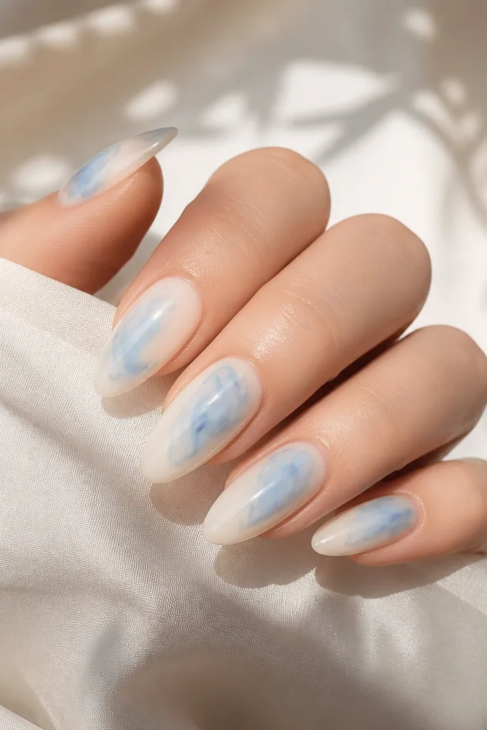 Almond nails with a sheer milky base and loose sky-blue watercolor swirls. The swirls fade at the edges like ink in water, with a soft, airy look. Glossy finish.