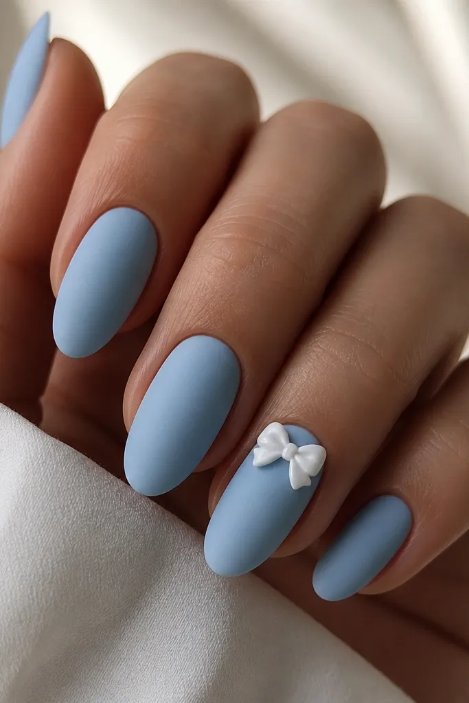 Almond nails with matte sky blue polish on all nails. On the ring finger, a glossy white bow sits near the cuticle with two curved loops and a tiny center knot. Other nails are plain matte.