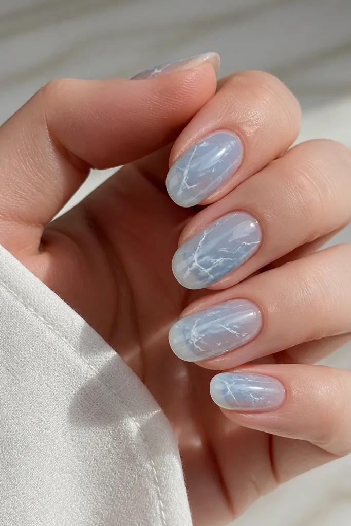 Medium oval nails with a transparent sky blue tint at the base, veined white marble lines branching diagonally across each nail. The marble looks like thin cracks with a glossy finish.
