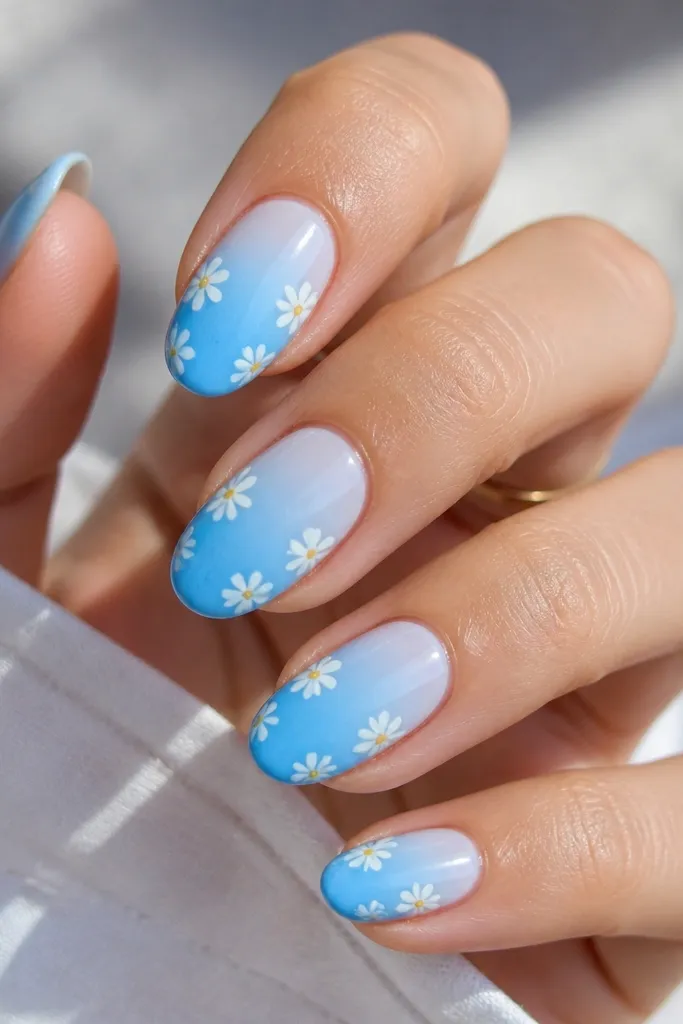 Medium oval nails with a sky blue gradient from light at the cuticle to deeper blue near the tips. Each nail has small white five-petal flowers spaced along the sides, plus tiny yellow dots in the center.