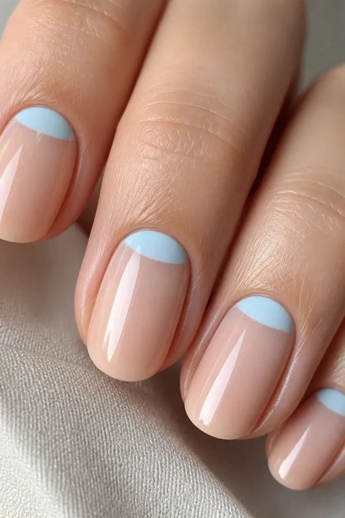 Four nails visible. The cuticle area has a sky blue half-moon shape under the skin line. The rest of each nail is sheer nude and glossy.