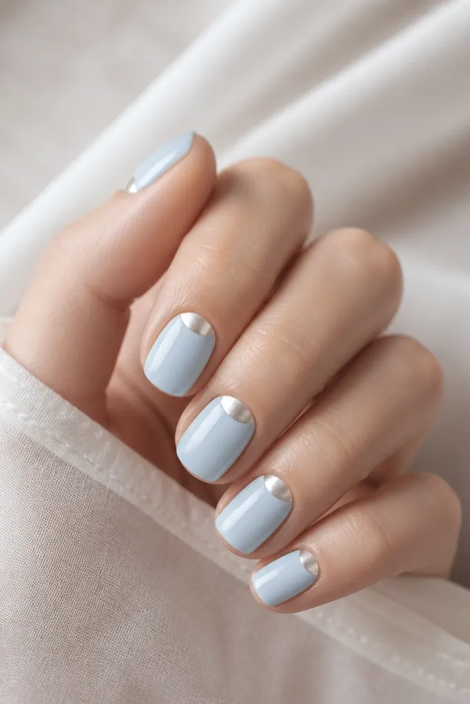 Short square nails painted powder blue with a glossy finish. Each nail has a pearl chrome half-moon at the cuticle area, like a reversed French.