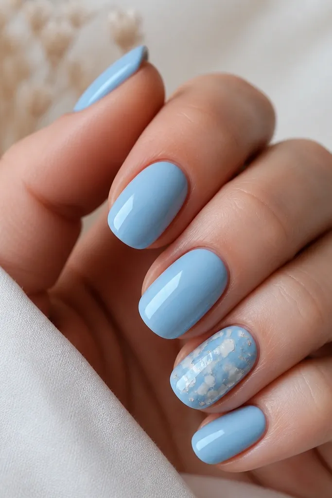 Most nails are glossy sky blue; one nail has delicate white floral foil fragments scattered near the center with a few tiny silver flecks.