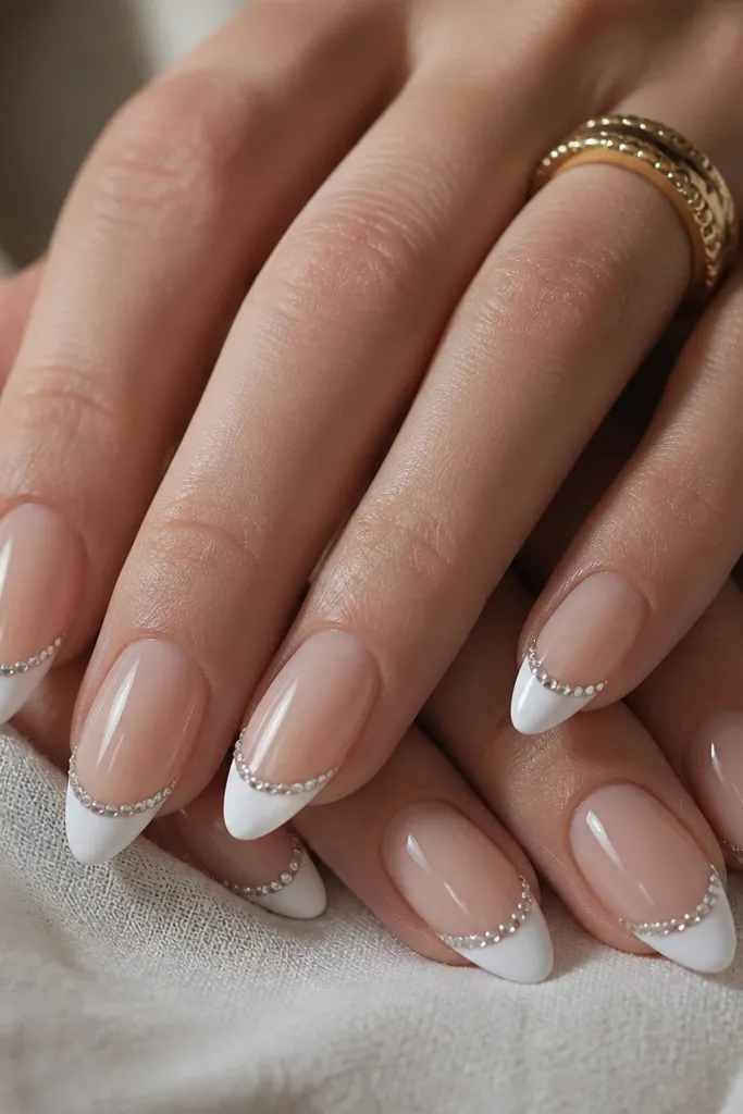 Almond nails with sheer nude base and bright white French tips. Along the top curve of the white tip there's a straight micro line of tiny clear rhinestones. Nails are glossy around the stones.