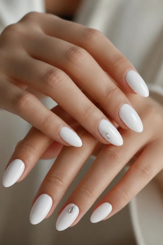 Glossy white almond nails with one small rhinestone set near the outer side of each ring finger, shaped like a teardrop. The rest of the nails are plain white.