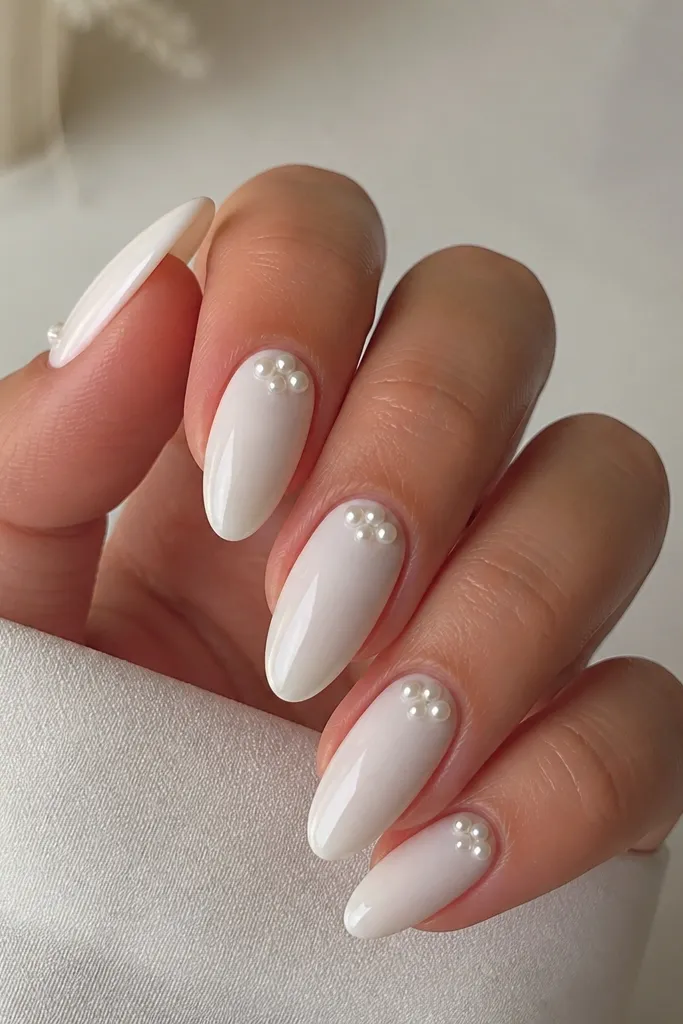 White almond nails with small pearl studs placed near the cuticle. Each nail has a tiny three-point cluster, and the rest is glossy white.