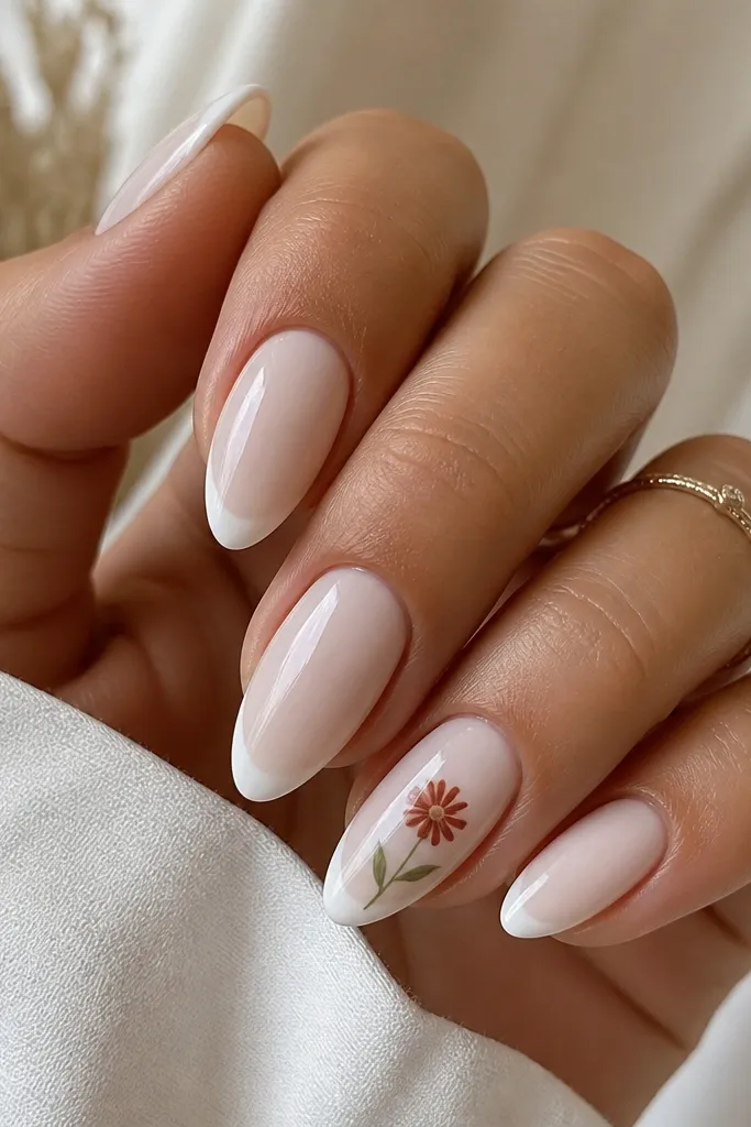 White almond nails with a subtle white French tip. On the ring finger only, a small terracotta daisy bloom sits near the cuticle with two green leaves and a thin stem line.