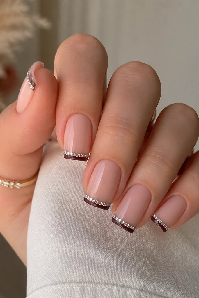 Square nails with a nude base and a thin espresso-brown French line. Along the very edge of the French, there is a single row of tiny rhinestones in a straight line.
