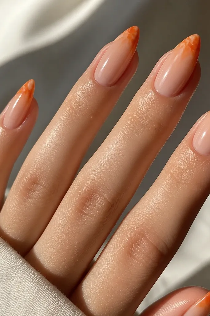 Almond nails with a sheer nude base. The tips show marbled orange and tangerine swirls like citrus peel, with a glossy top coat reflecting light.